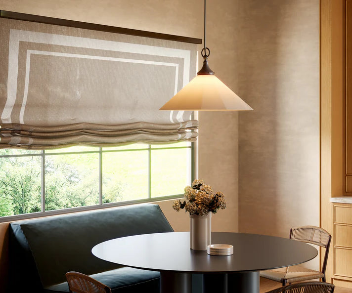 Shows a dining room with a round hanging light over a round table with chairs to the side and a bench seat against a window. The large window behind the bench is partially covered by blinds.