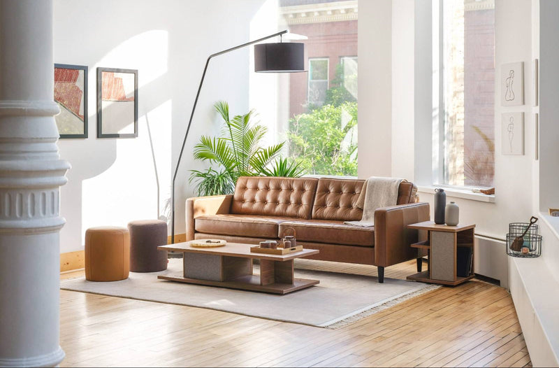 A brown tufted leather sofa in a bright, modern living room with light wood floors, a white rug, a wooden coffee table, and wooden side table in walnut.