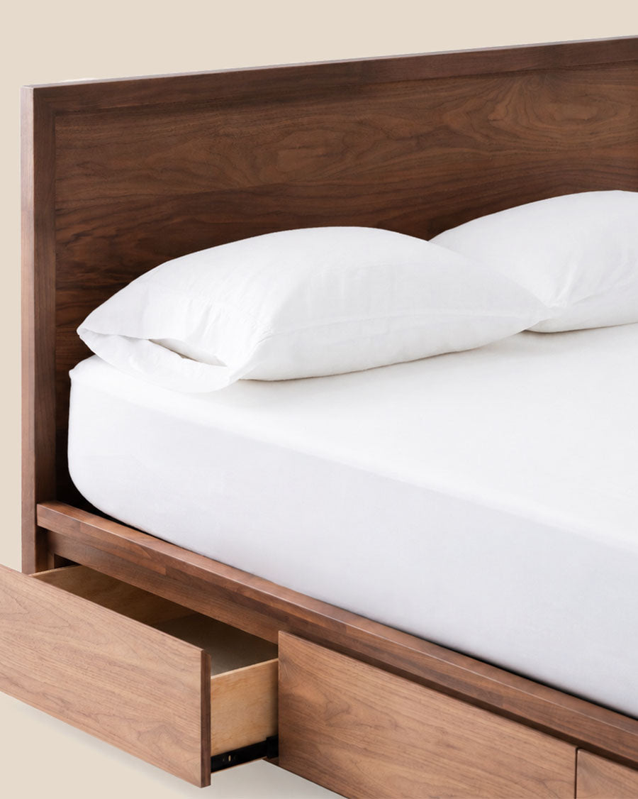 Un gros plan d'un cadre de lit moderne avec une tête de lit en bois de noyer foncé et des tiroirs de rangement assortis, habillé de literie blanche.