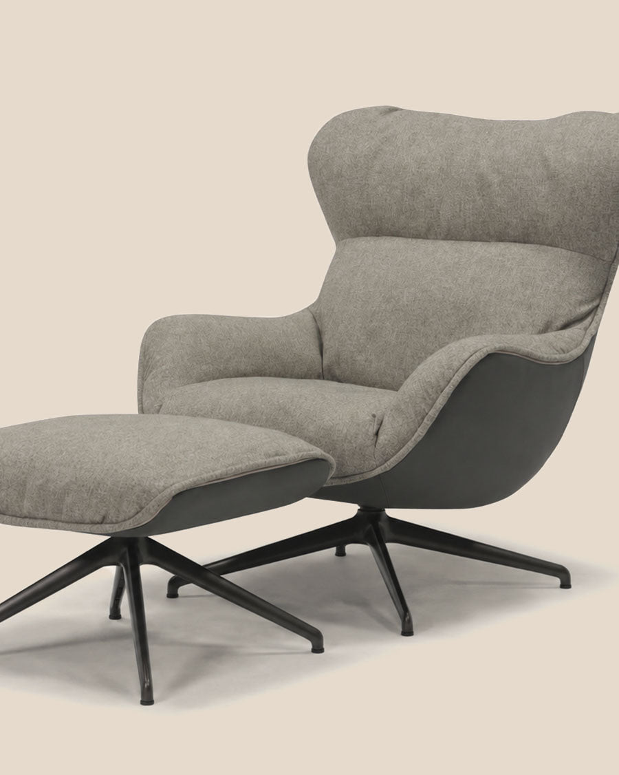 Un fauteuil de salon moderne rembourré avec pouf assorti en tissu gris clair et panneaux latéraux gris foncé, sur pattes pivotantes noires, photographié sur fond beige clair uni.