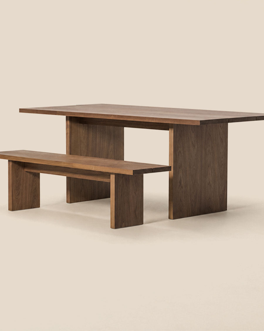 Une table à manger en bois brun foncé avec un banc assorti, présentée dans un décor de studio épuré sur fond blanc cassé chaleureux.
