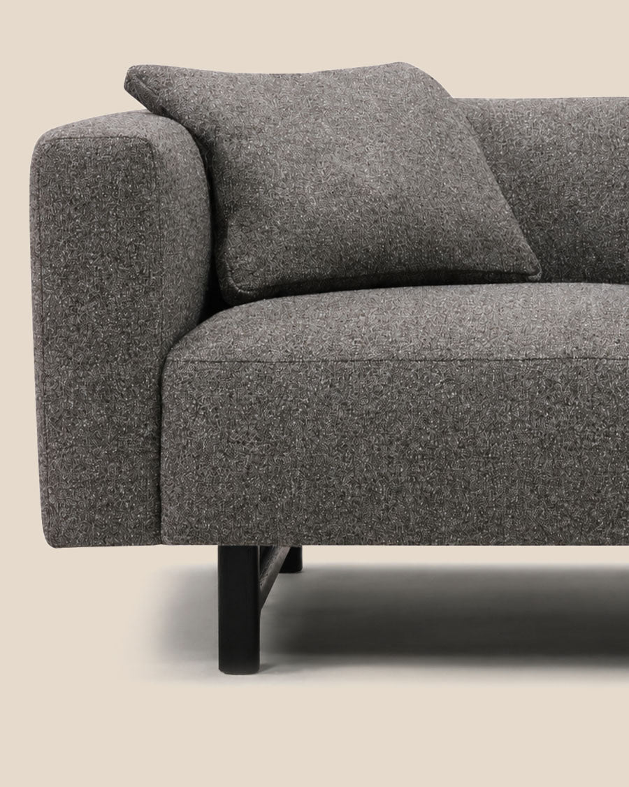 Gros plan d'un coin de canapé moderne gris rembourré avec un coussin décoratif assorti et une patte noire, présenté sur fond beige clair.