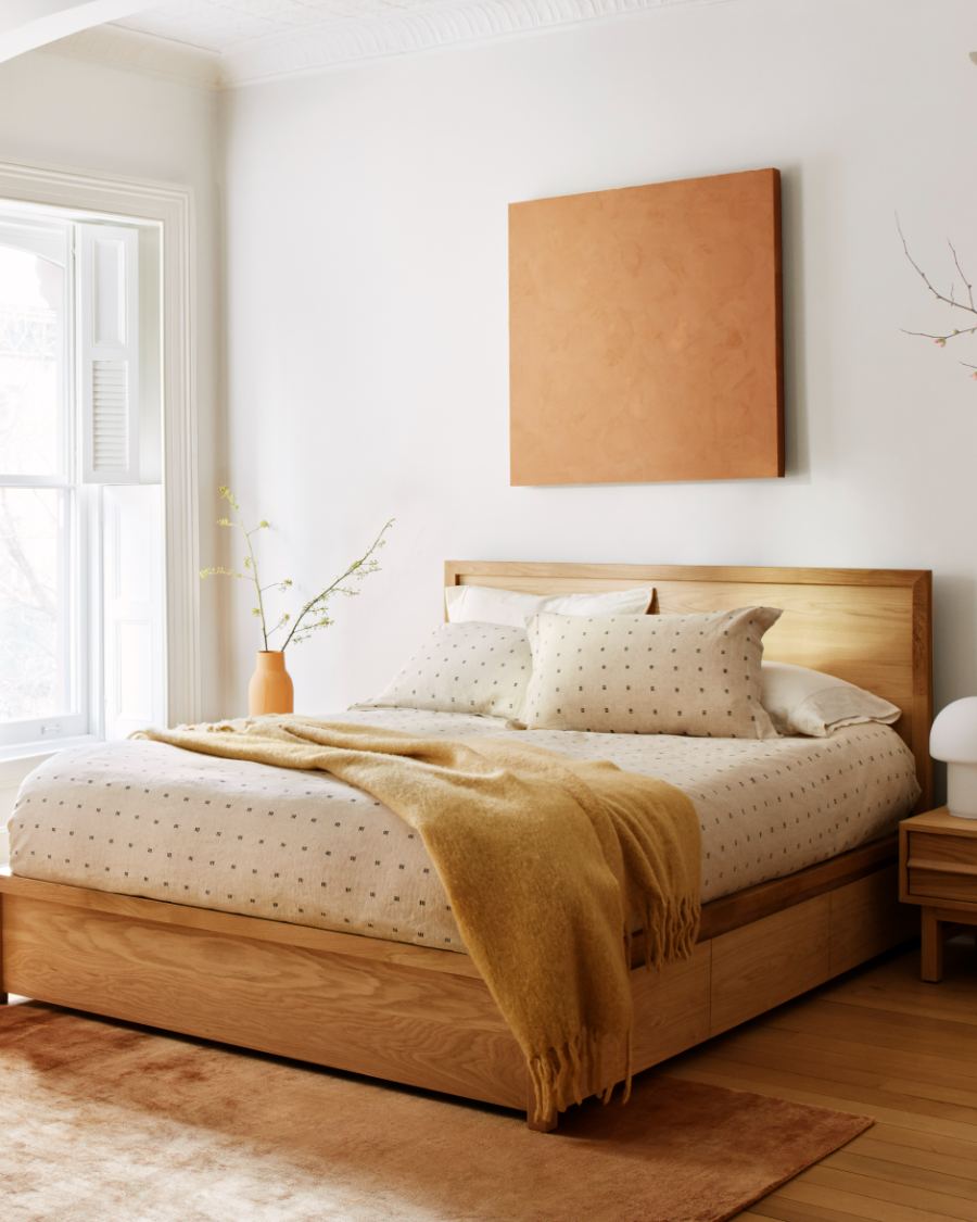 Une chambre minimaliste avec un lit plateforme en bois de chêne clair, une literie crème à motifs et une couverture jetée moutarde. Une table de chevet en bois assortie est placée à droite, et une toile carrée en terre cuite texturée est accrochée au mur blanc au-dessus de la tête de lit.