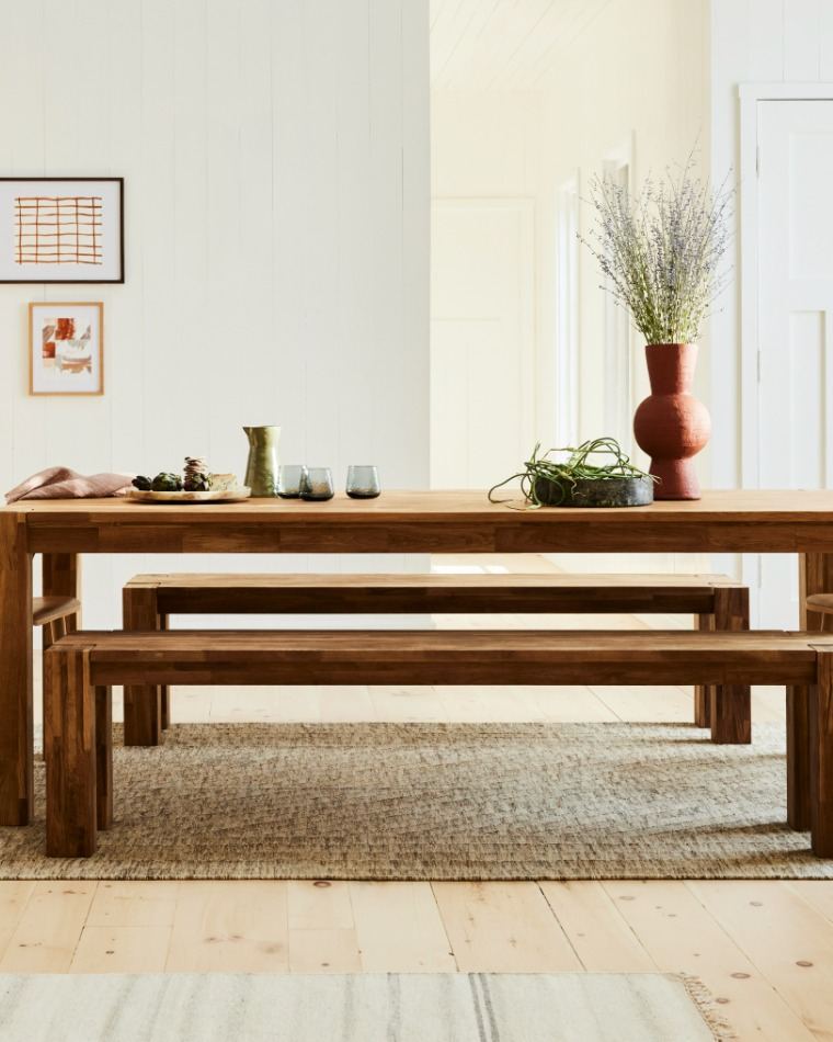 Une table à manger rustique en bois avec des bancs assortis dans une salle à manger lumineuse et moderne. La table et les bancs sont en bois brun moyen chaleureux, posés sur un tapis tissé texturé de couleur beige.