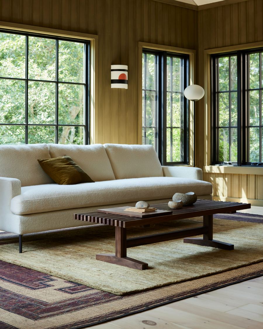 Un canapé rembourré crème dans un salon rustique aux murs lambrissés de bois, avec de grandes fenêtres à cadre noir, une table basse en bois foncé et un tapis à motifs.