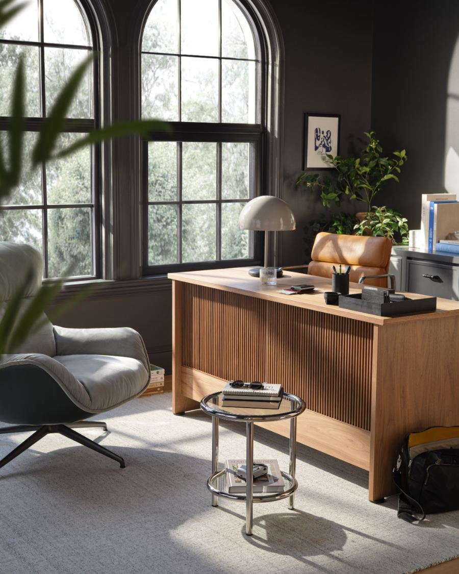 Une scène de bureau à domicile moderne avec un bureau en bois de chêne clair à façade cannelée, agencé avec une lampe de bureau blanche, une petite table d'appoint ronde en verre, un fauteuil gris et une chaise en cuir, dans une pièce aux murs gris foncé et aux hautes fenêtres arquées.