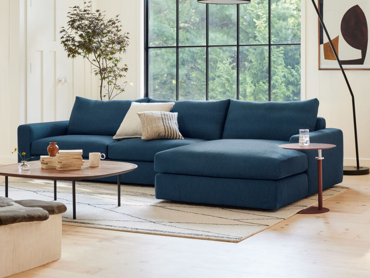 Un canapé sectionnel bleu moderne avec méridienne dans un salon contemporain et lumineux. Le canapé est agencé avec des coussins décoratifs neutres et posé sur un tapis de couleur claire, à côté d'une table basse ronde et d'une petite table d'appoint.