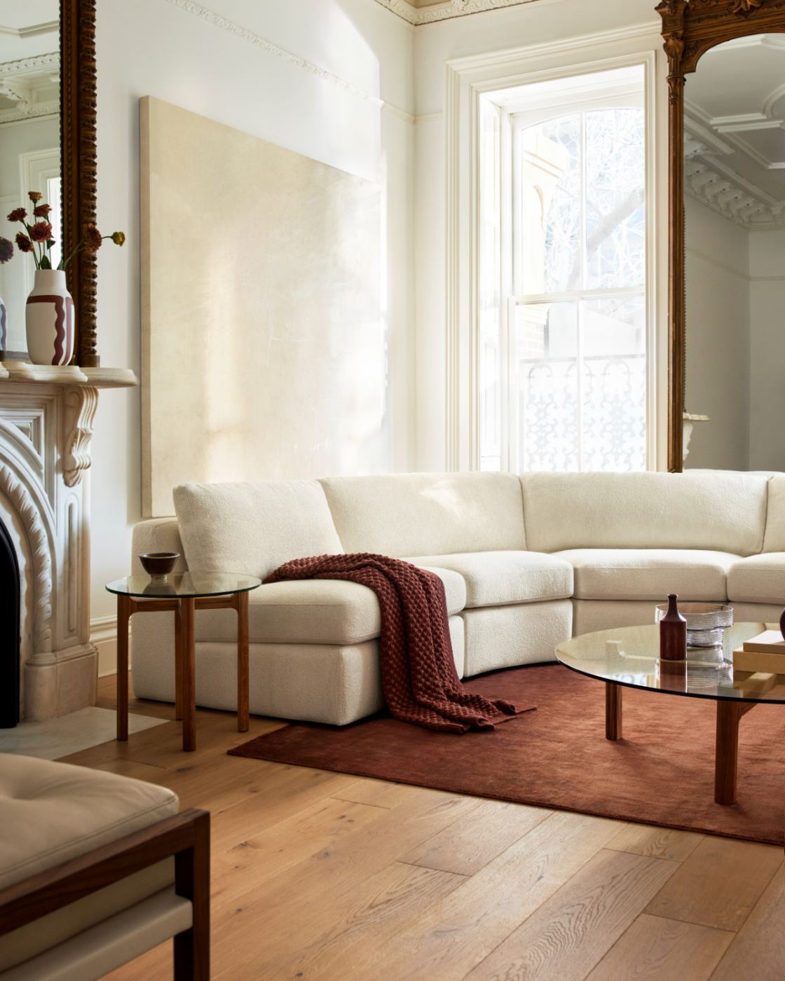 Un canapé sectionnel blanc moderne et incurvé dans un salon lumineux et élégant, avec des planchers en bois clair, un tapis rouille et une table basse à plateau en verre.