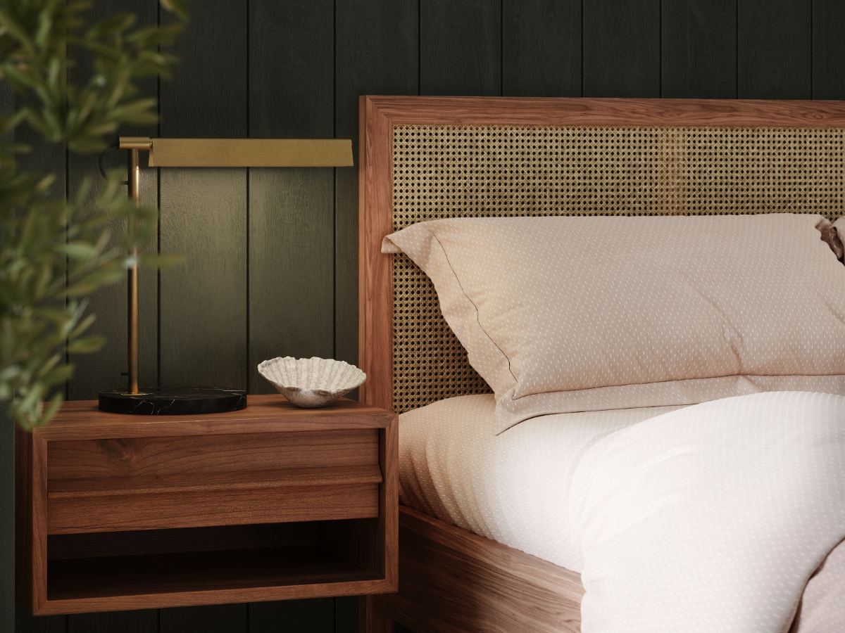 Un gros plan d'un cadre de lit en bois avec une tête de lit en cannage tressé, habillé de literie crème, à côté d'une table de chevet en bois assortie portant une lampe de table en laiton et un petit plat blanc, sur fond de mur lambrissé foncé.